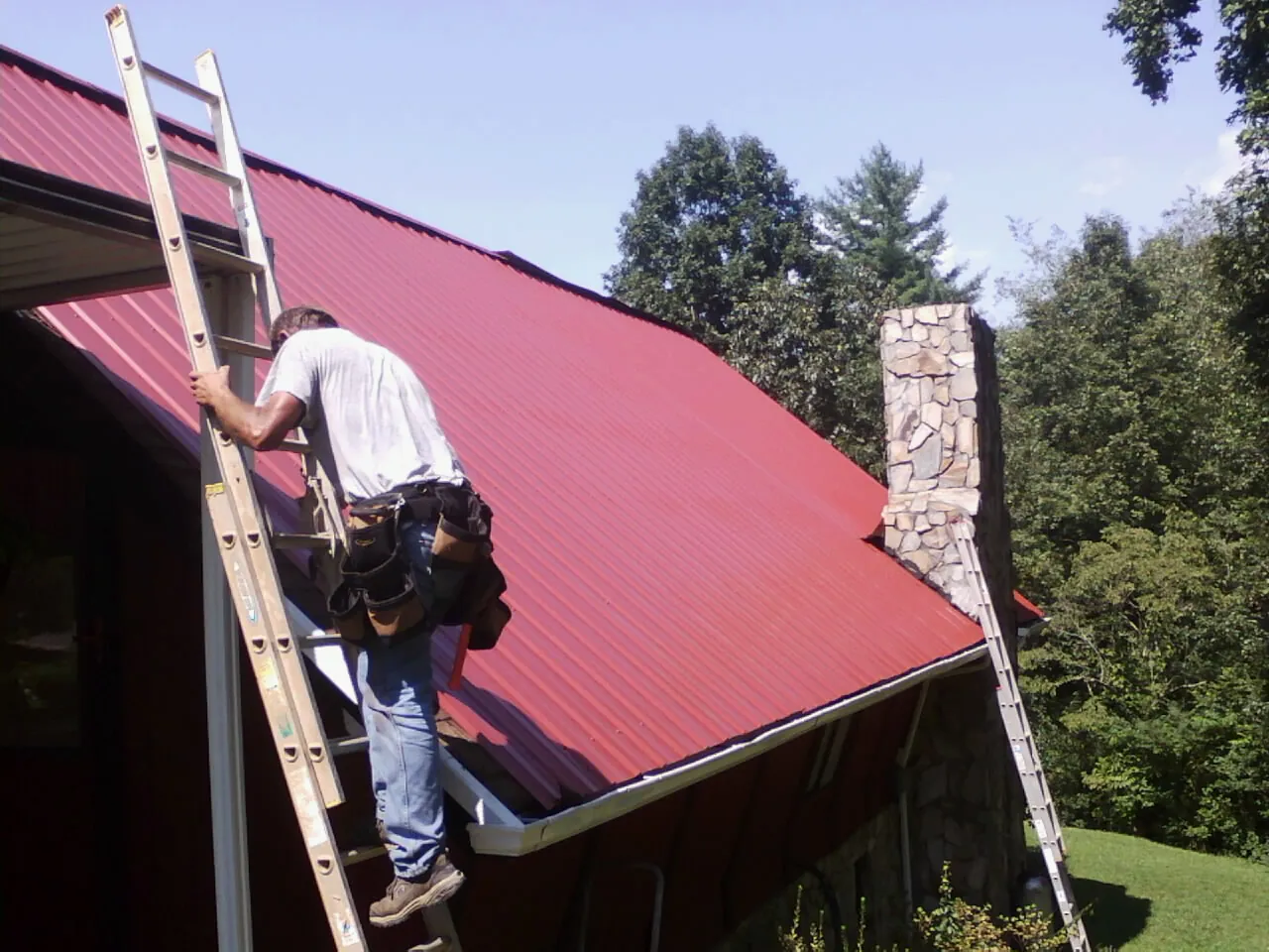 Roofer on a ladder beside a red metal roof and stone chimney — Soffit & Fascia Repair by Roofing Brothers in Cos Cob