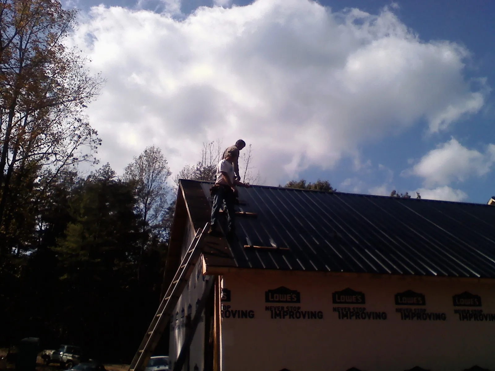 Roofer fastening dark metal panels high against a cloudy sky, from a Roof Inspection job in Cos Cob